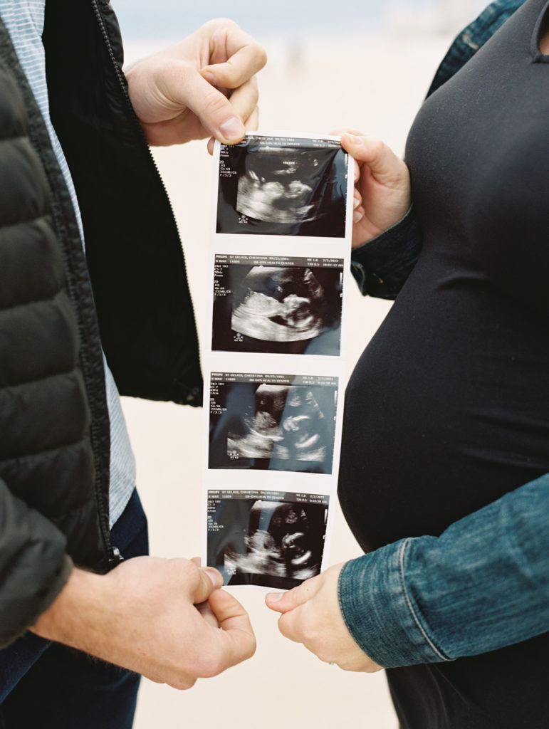 a couple holding four ultrasound photos of new baby in front of the woman's pregnant belly