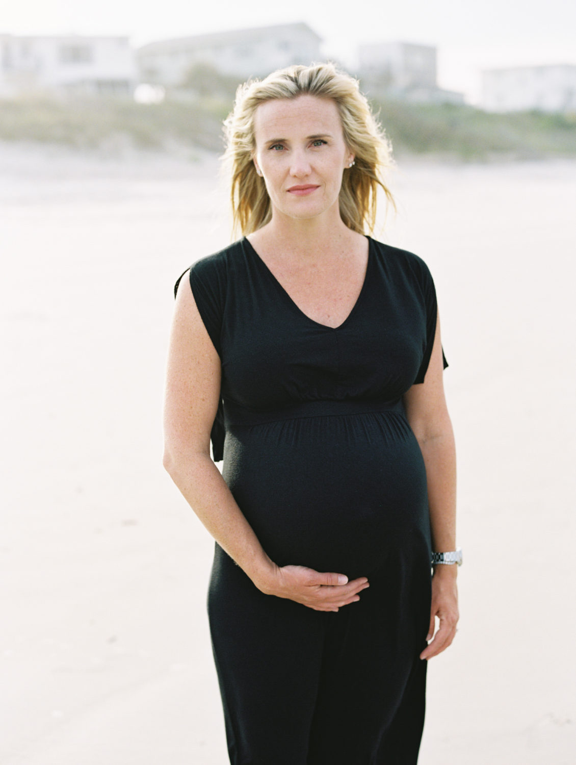 woman with blonde hair and blue eyes in a black maxi dress looking at the camera with a serious expression and holding her pregnant belly with one hand