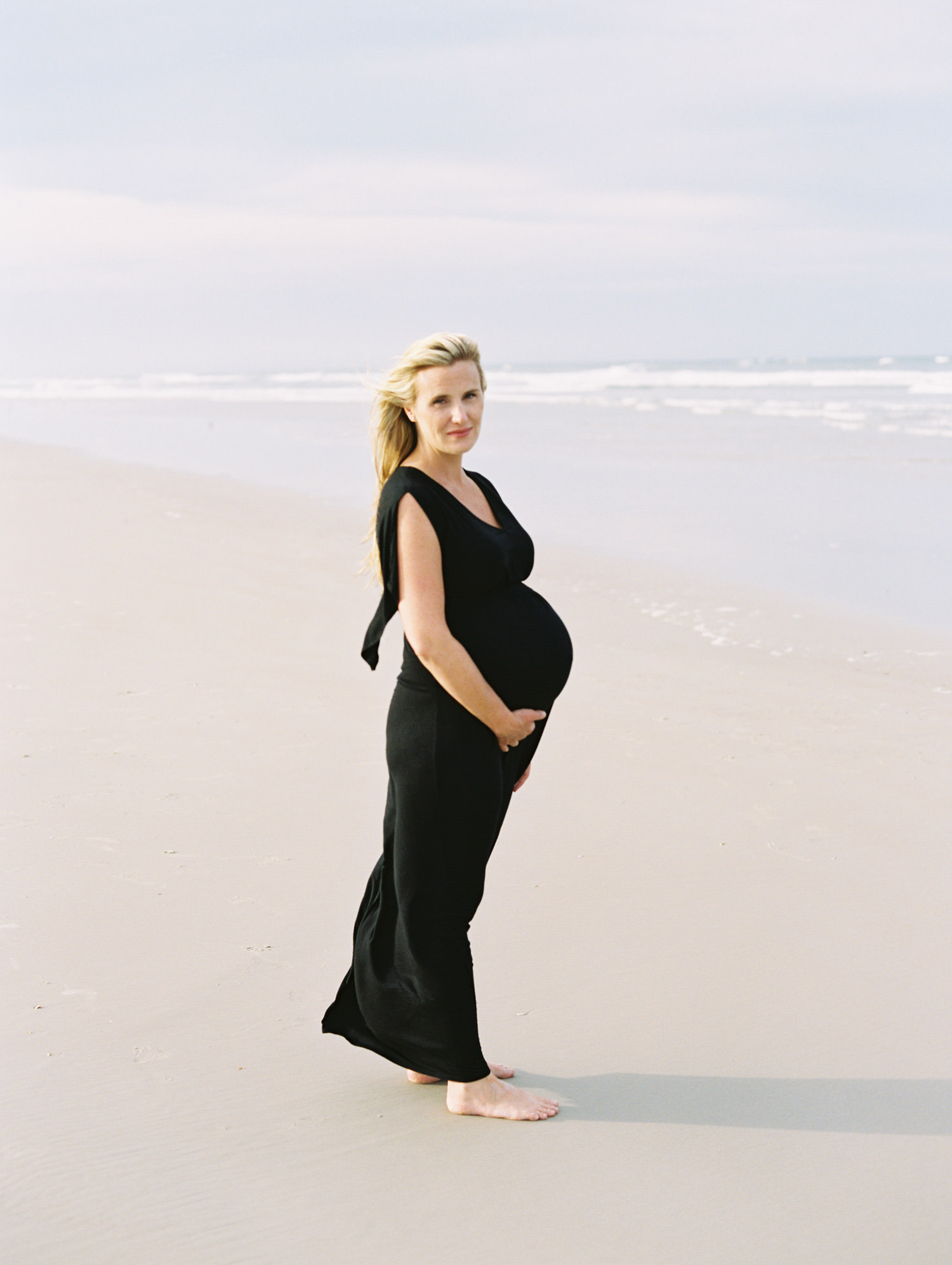 pregnant women standing in profile in a black maxi dress and looking at the camera with the ocean behind her