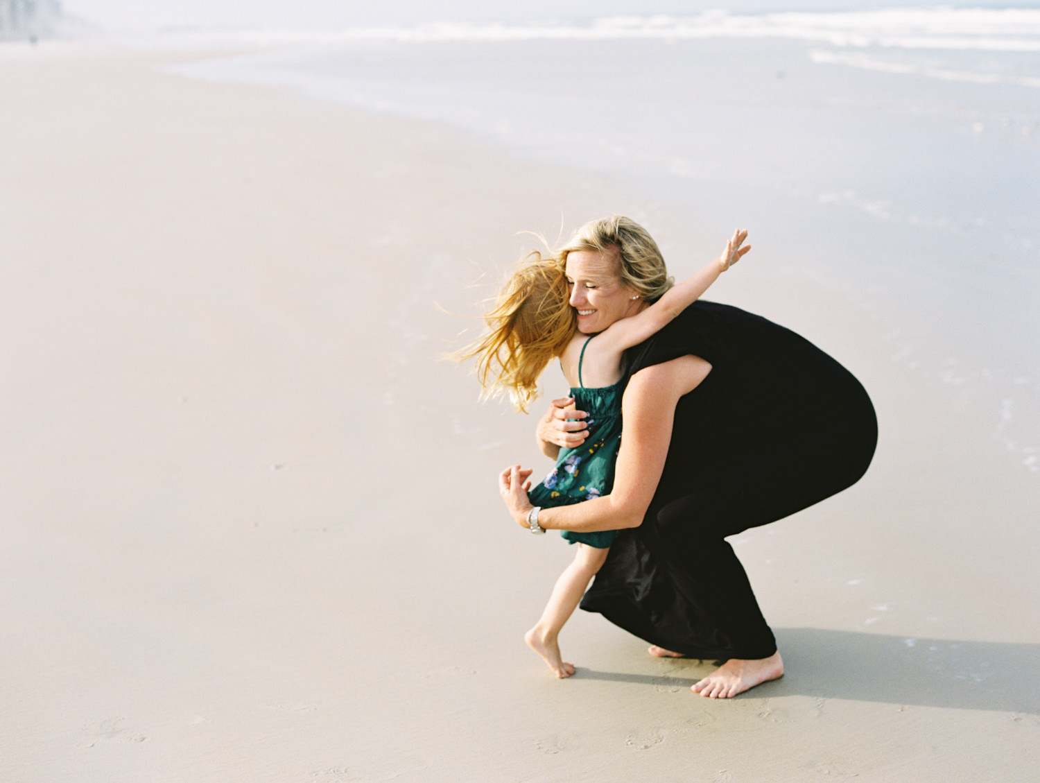 toddler in a green dress and red hair flings her arms around her mother's neck as she bends down to pick her up.