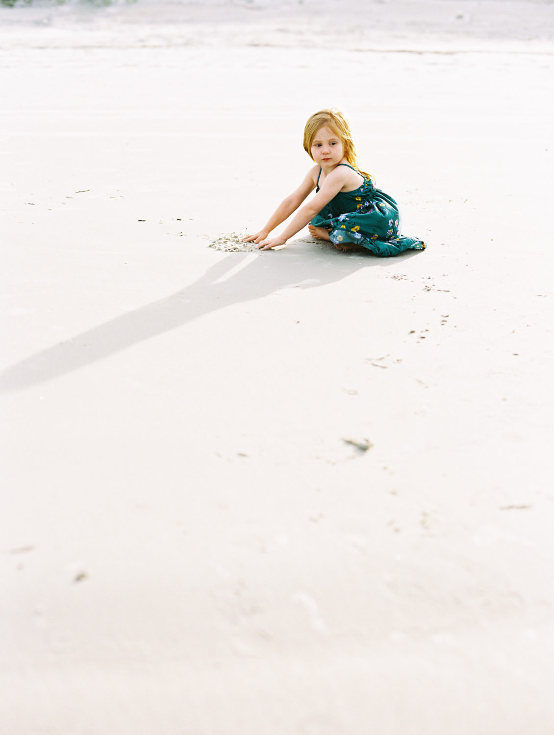 little girl in green sundress and red hair sits quietly on the beach rubbing her hands in the sand as the sun casts her shadow