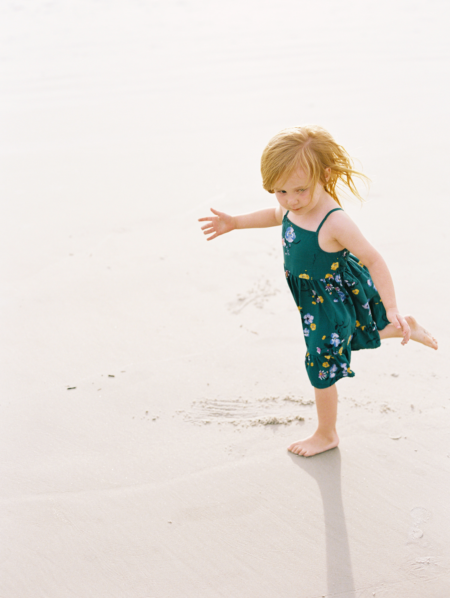 red haired toddler girl in green floral dress looks down and runs barefoot on the beach