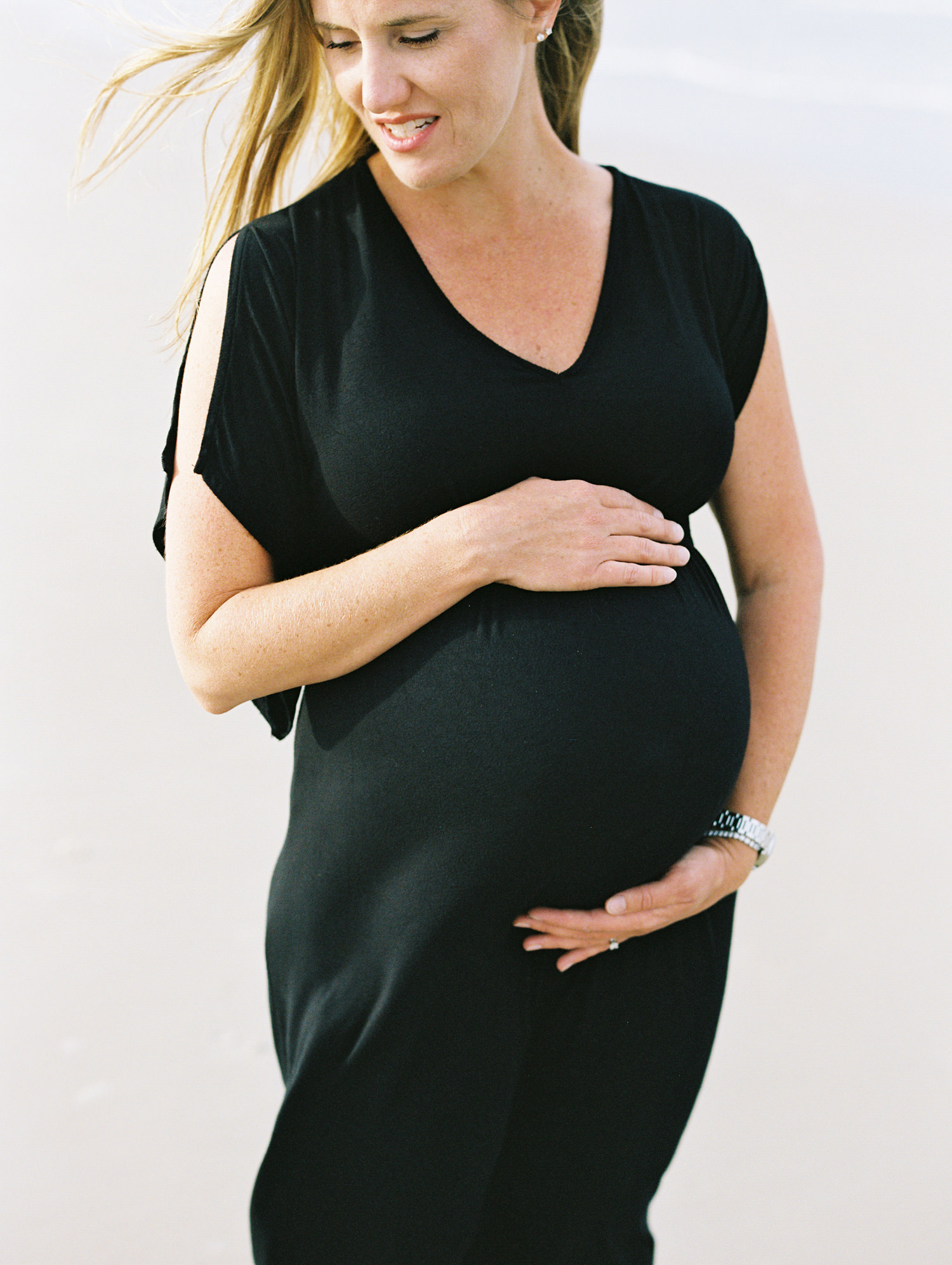 woman in a black v neck dress and long blonde hair holds her pregnant belly while looking down at her toddler