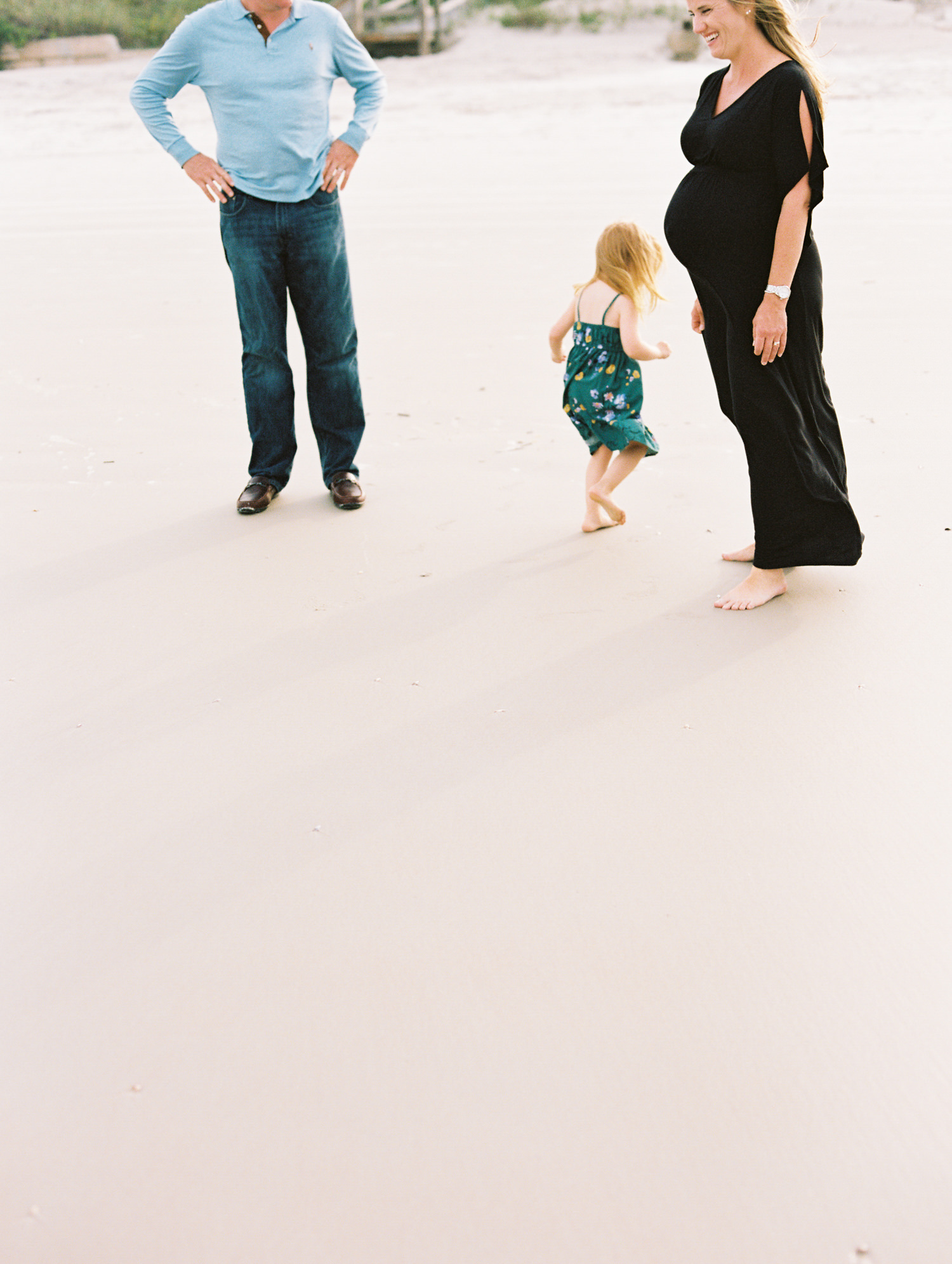 pregnant woman with long blonde hair in a long black dress laughing on the beach while her daughter plays