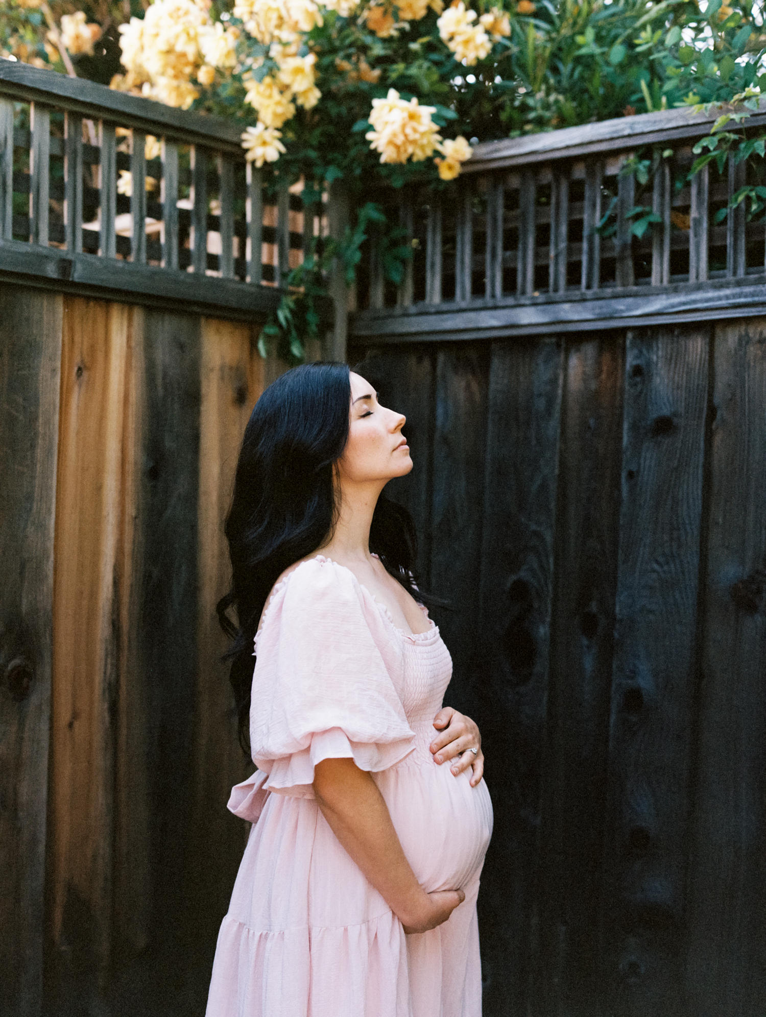 pregnant woman in blush dress standing in front dark fence with her eyes closed and face up to the sky