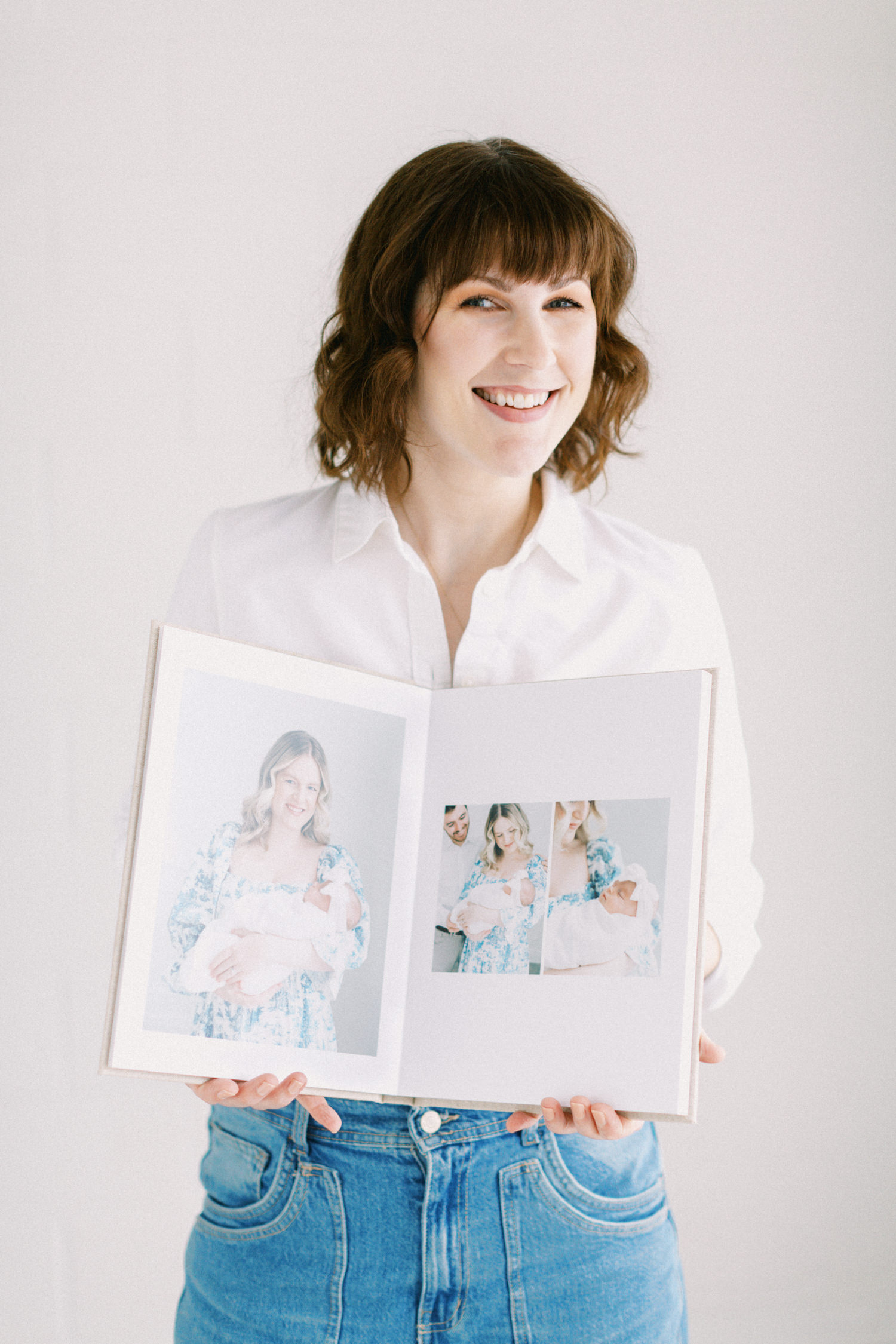Toronto Newborn Photographer Emily Anderson smiling at the camera wearing a white button up shirt with jeans holding a fine art newborn album