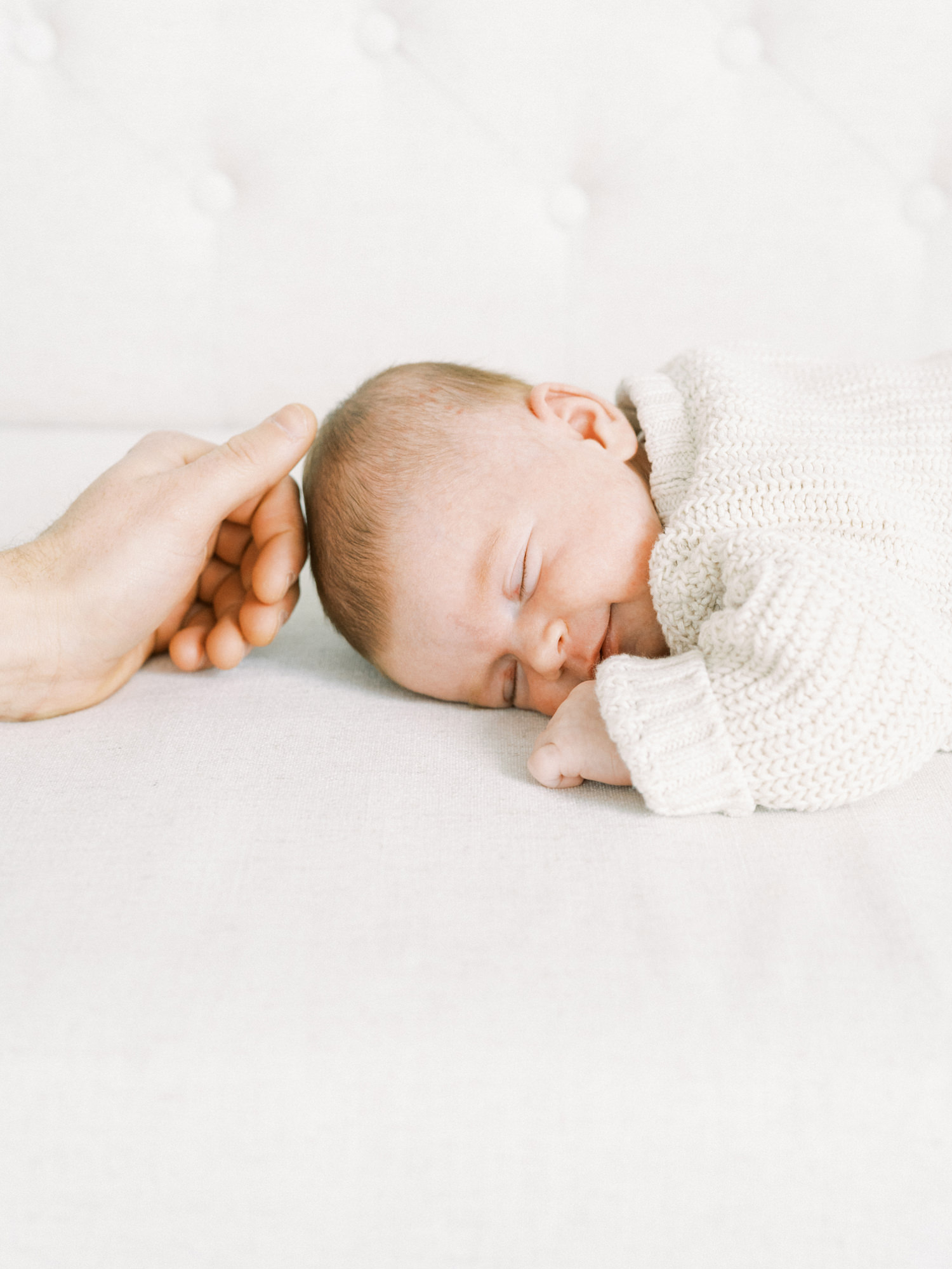 sleeping newborn on white couch wearing white sweater smiling as father strokes their head