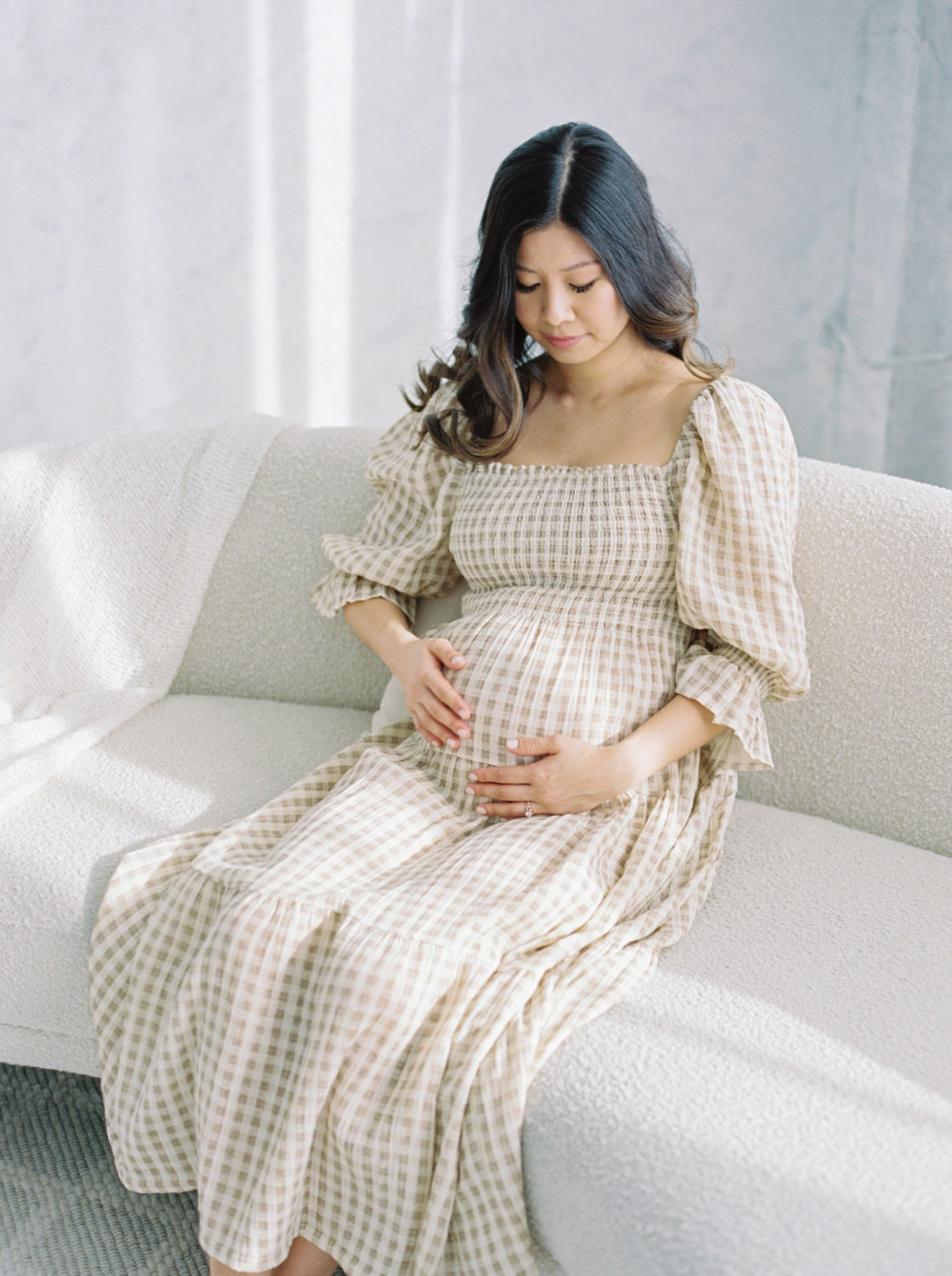 pregnant woman sitting down on a white couch holding her belly in a light gray photo studio