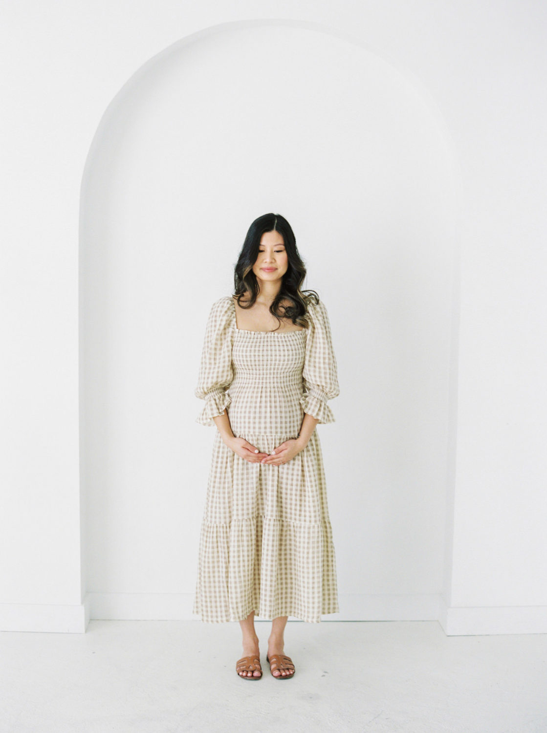 Pregnant woman in a three quarter sleeved tan and white dress and brown sandals in front of a white wall with a simple arch