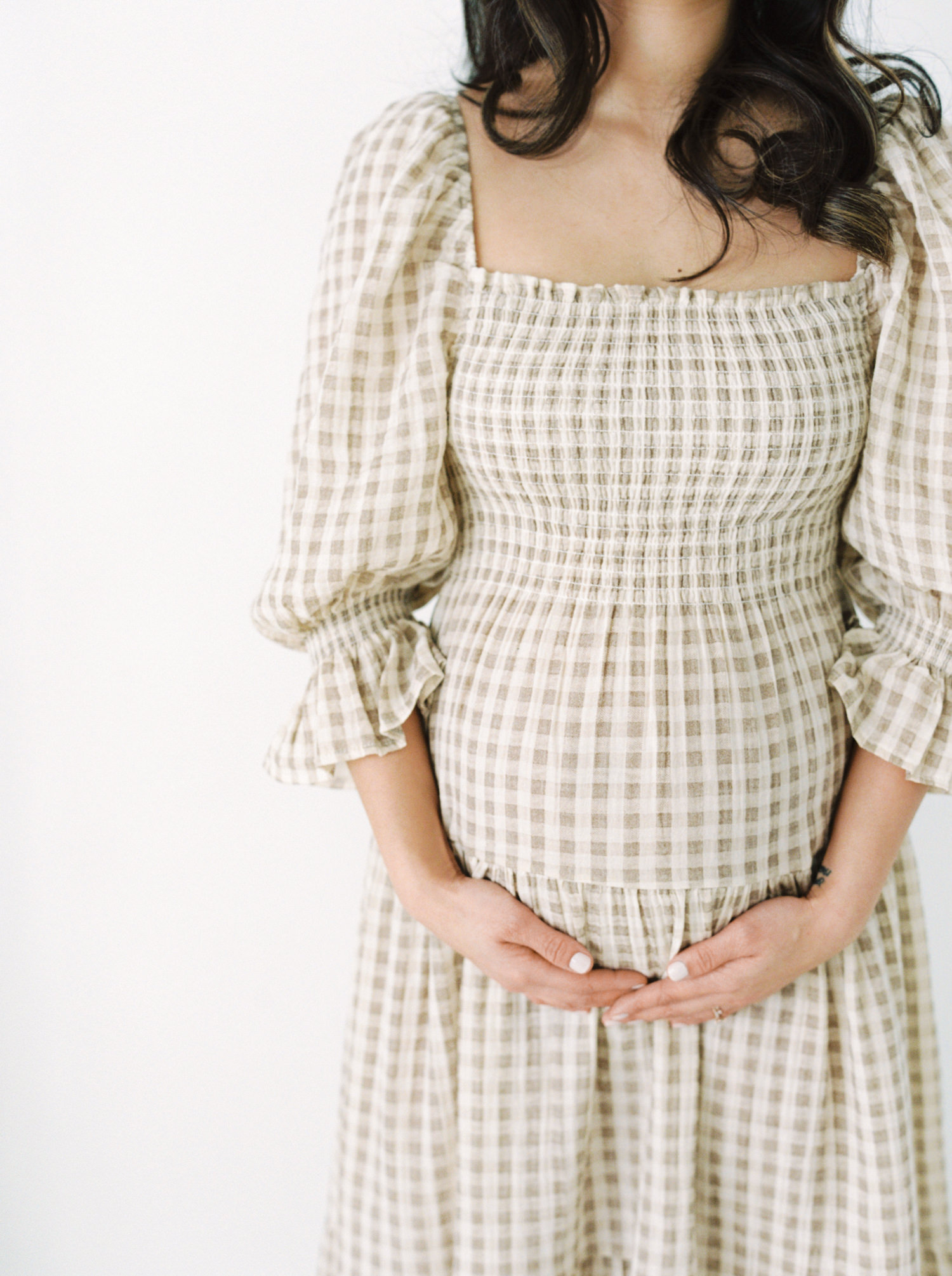 Expecting mother shown from the shoulders down in a smocked brown and white maternity dress in a markham photo studio