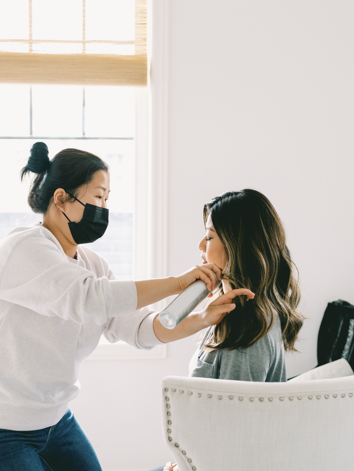 mom with long black hair sitting in a white chair having her hair and makeup styled by a woman in a white shirt and a black face mask