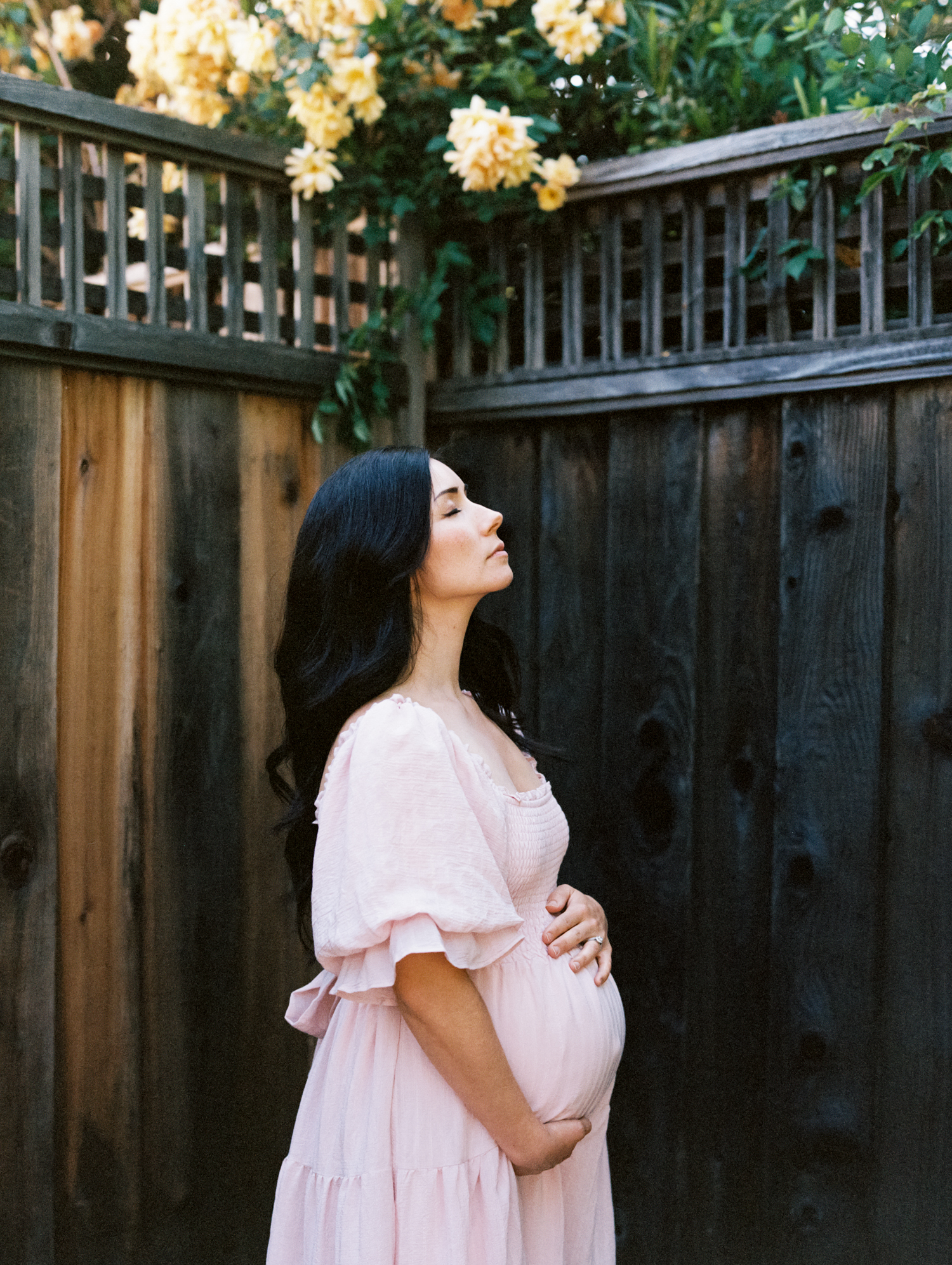 pregnant woman with long black hair in light pink dress standing in front of a dark fence with roses and closing her eyes