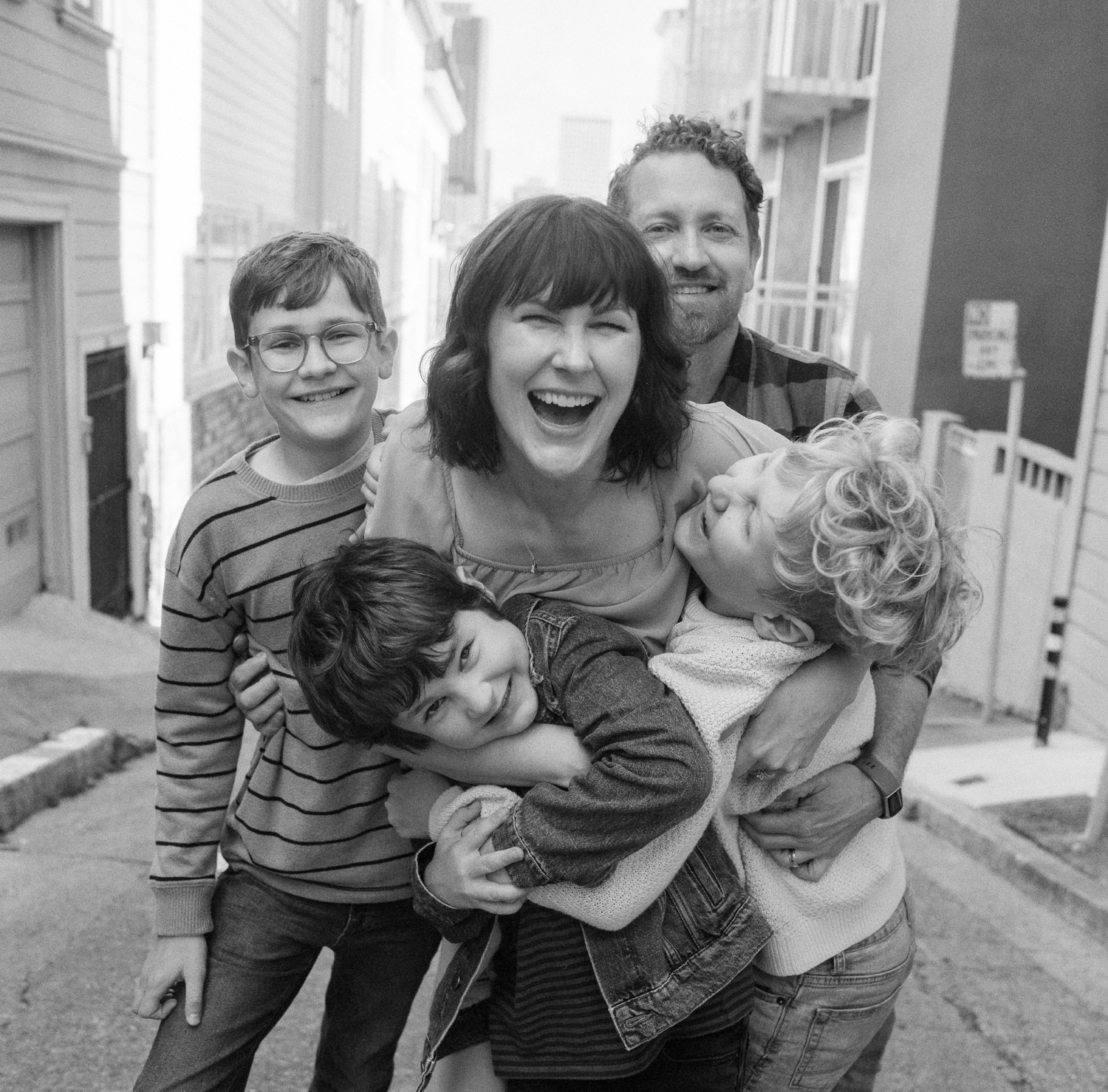 black and white image of photographer e m anderson laughing at the camera as she hugs her three boys with husband smiling in the background