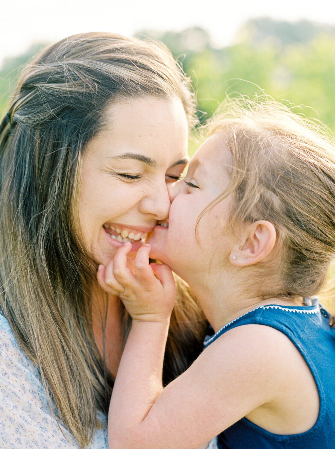Mom with long brown hair laughing while her toddler daughter kisses here cheek on a sunny summmer morning