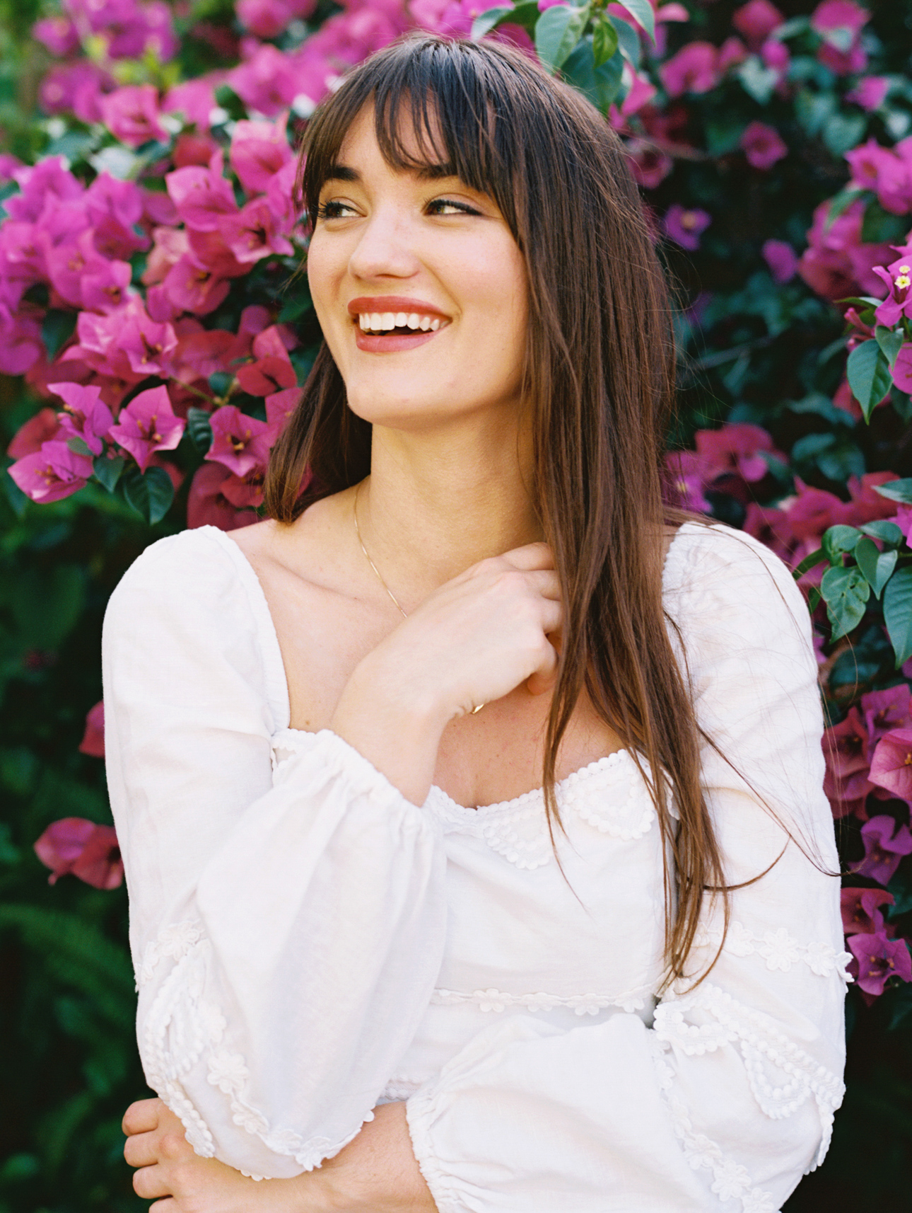 Woman with long brown hair smiling in a white dress in front of a wall of bougainvillea