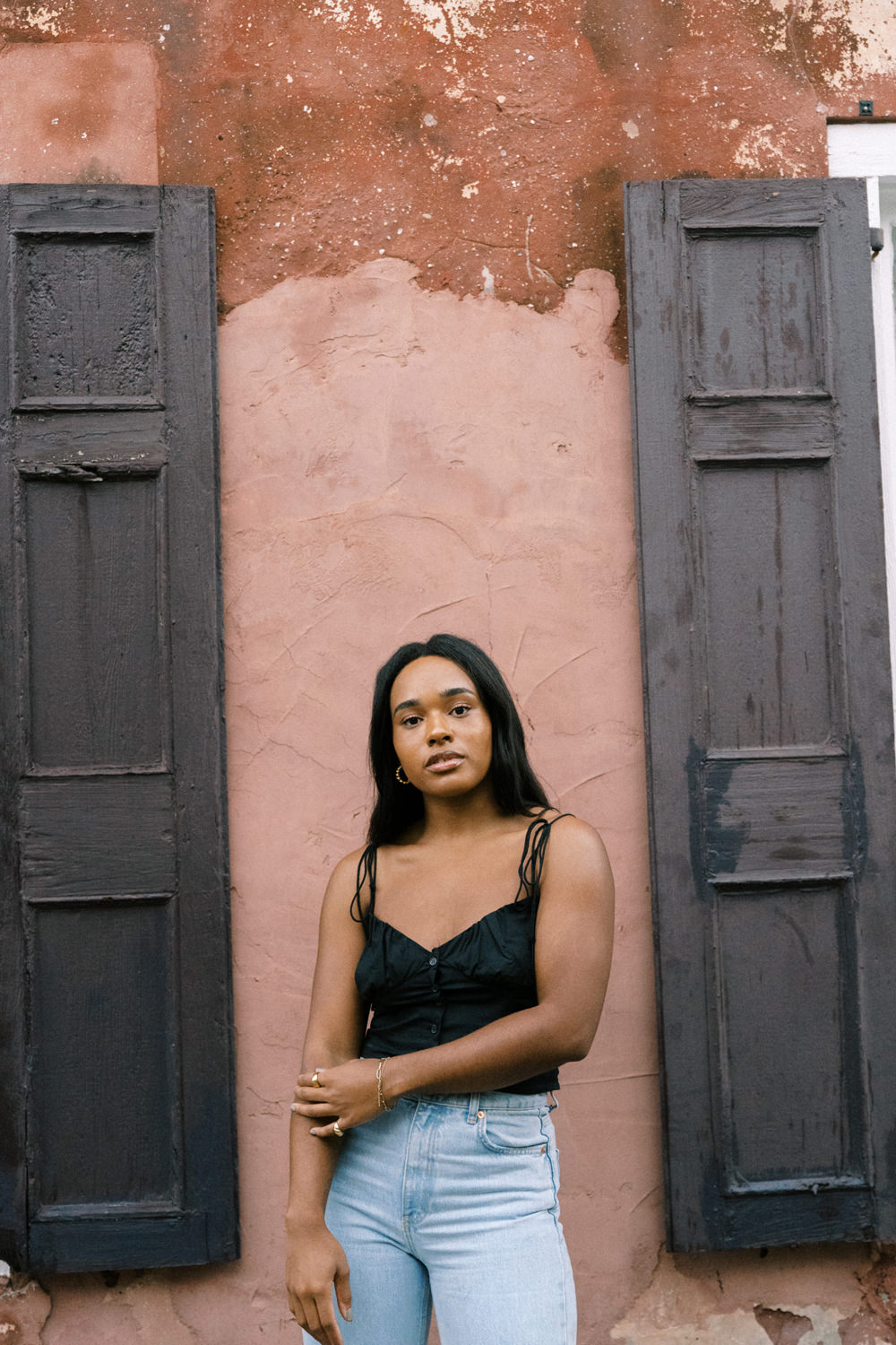 black woman with long black hair in a black tank top and jeans standing in front of a terracotta color wall with brown shutters