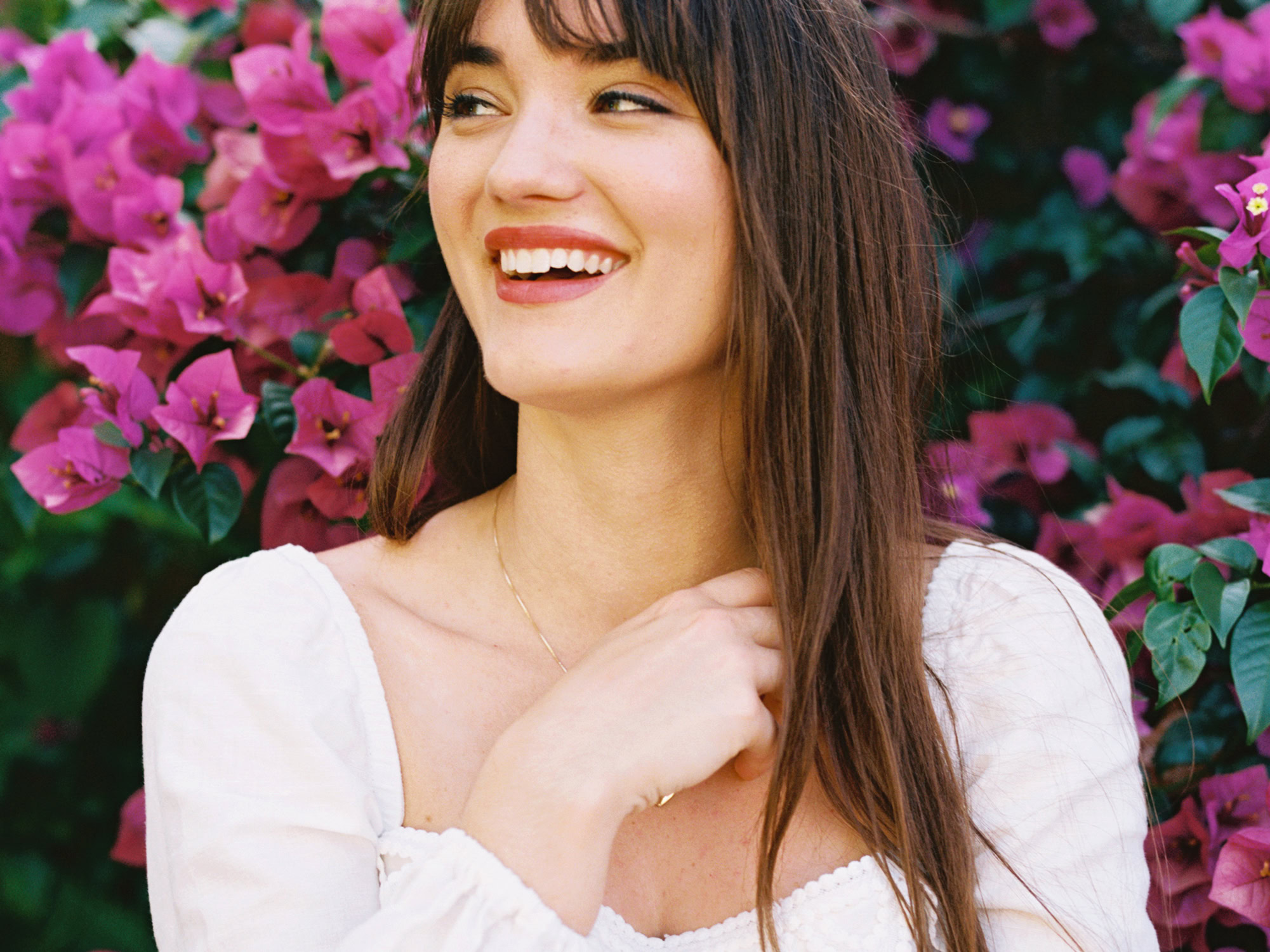 white woman with long brown hair and bangs, wearing red lipstick and a  white dress looking away from the camera and smiling in front of pink flowers