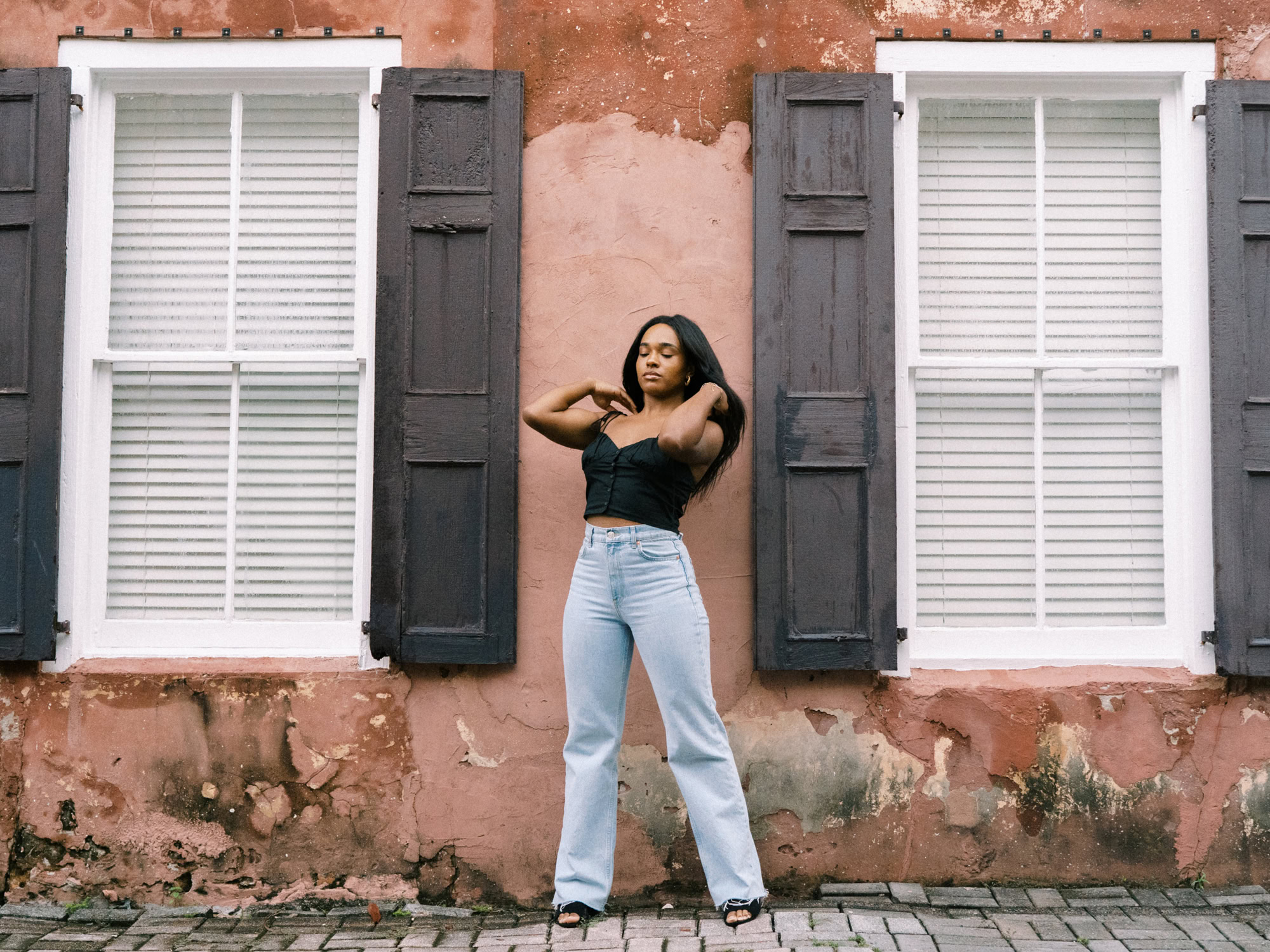 black woman with long black hair wearing a black tank top and jeans standing in front of a rose color wall with brown shutters