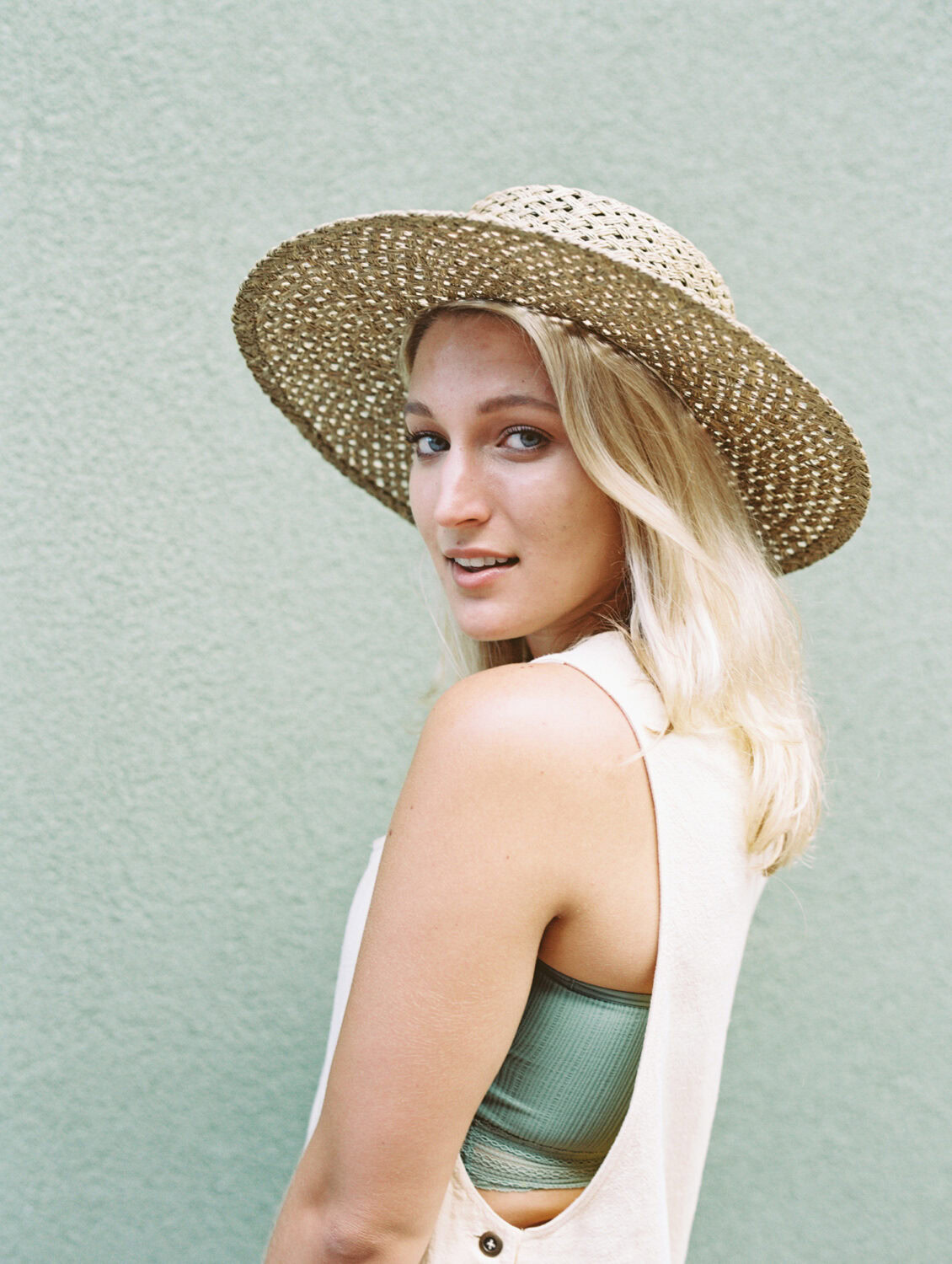 blonde woman in straw hat and white overalls with green top looking over her shoulder in front of a green wall