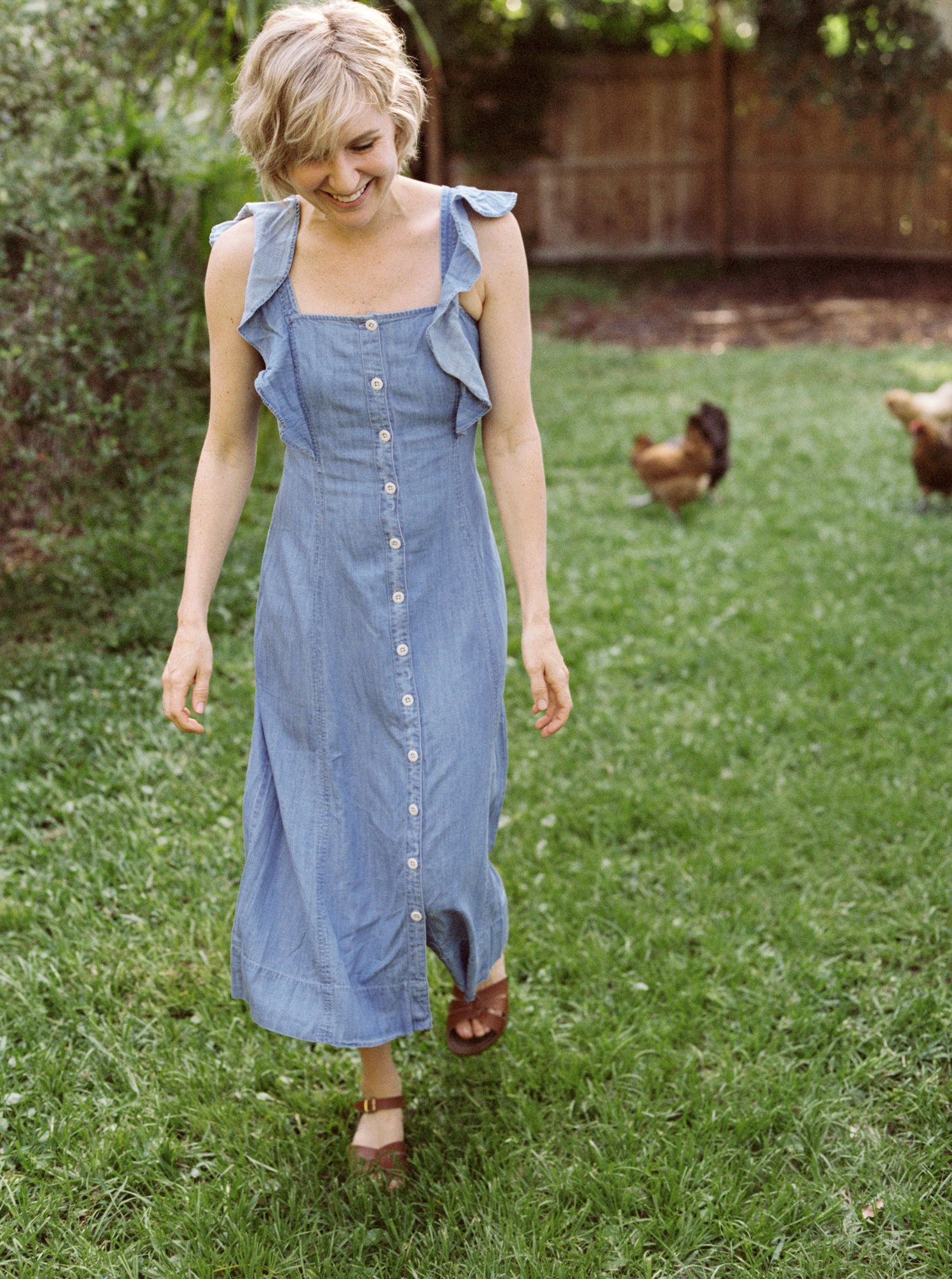 white woman with short blonde hair in a light blue dress walking in the grass with chickens behind her