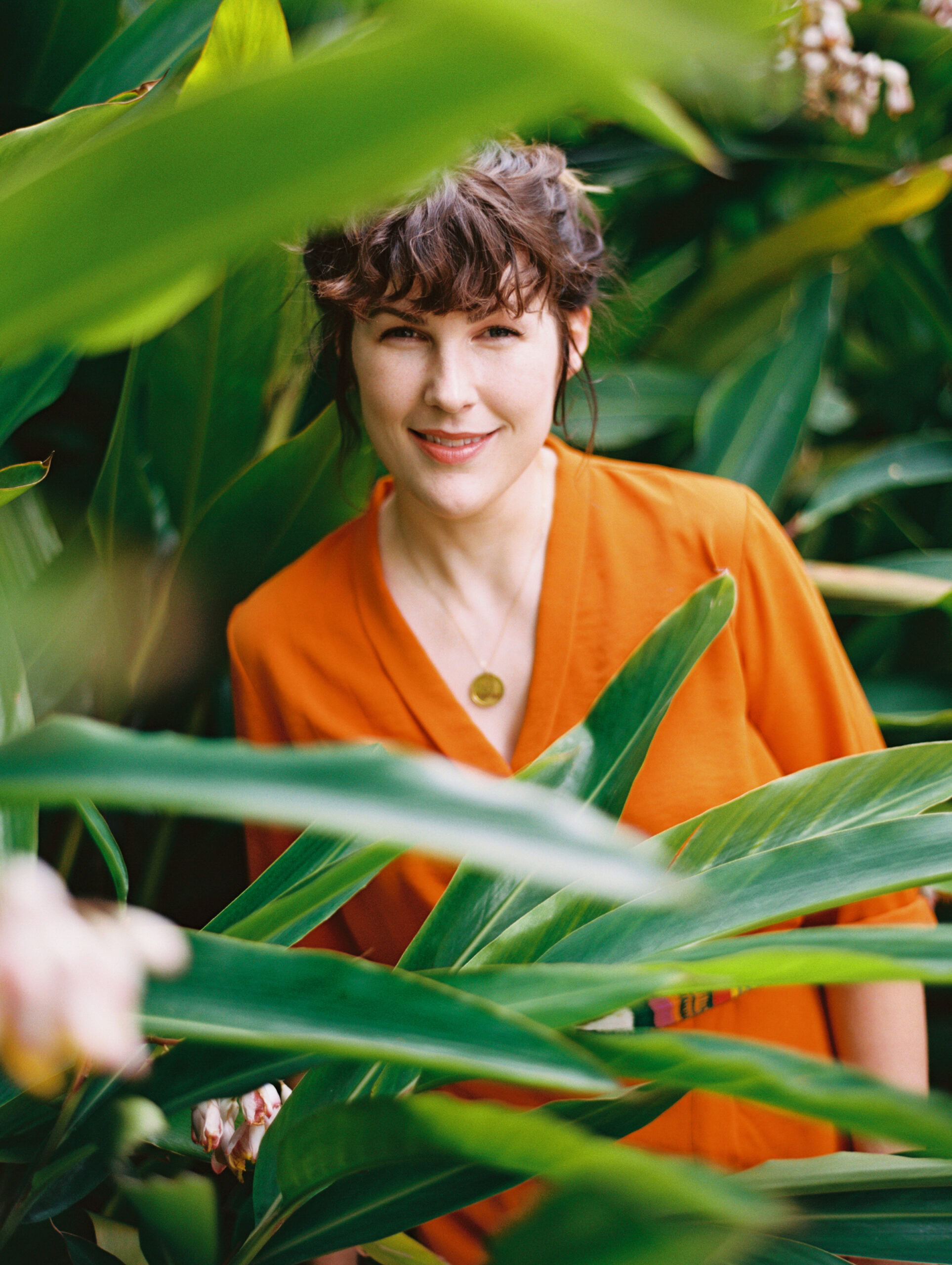 brand photographer emily anderson in an orange dress standing in tropical plants smiling at the camera