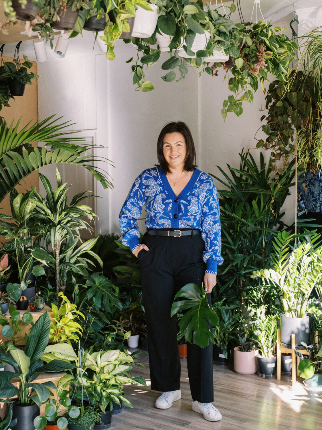 woman with brown hair and a blue sweater standing surrounded by large indoor plants