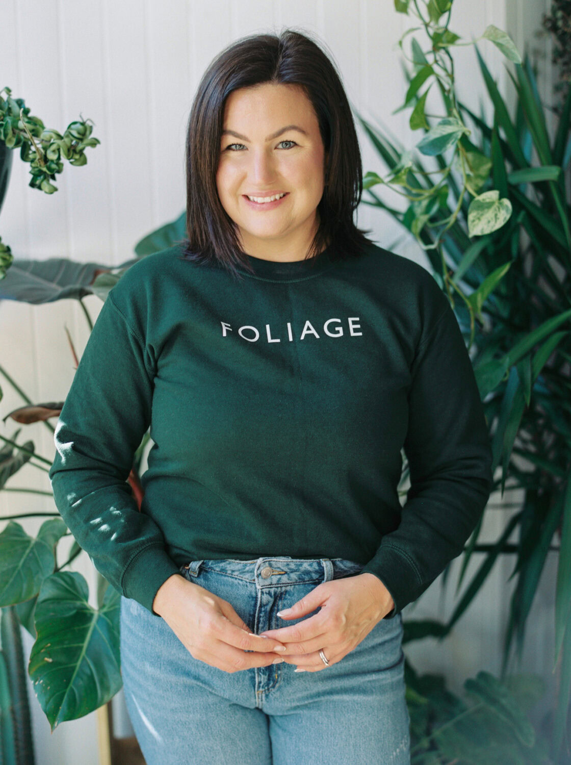 woman with brown shoulder length hair in green shirt and jeans smiling at the camera in a houseplant store