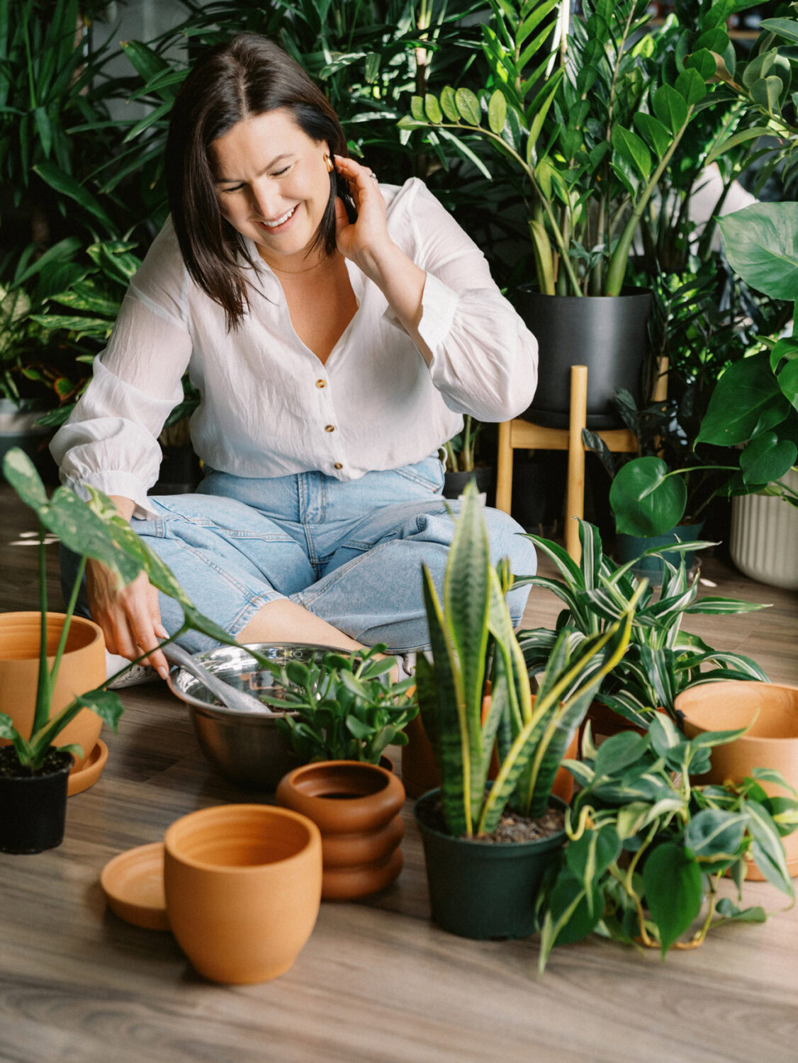 woman in white shirt and jeans sitting on the floor and tucking her hair behind her ear with pots and plants around her