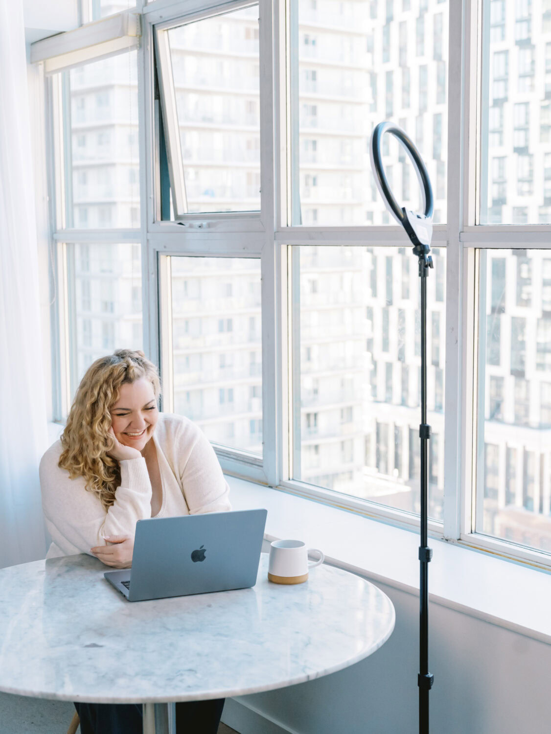 woman with long blonde curly hair in a white sweater sitting at a small table by a big windowi n a Toronto high rise and smiling down at her laptop