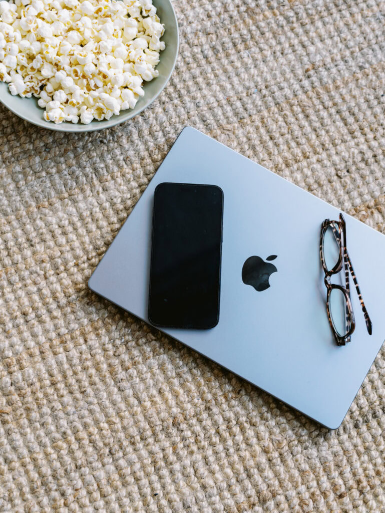 apple laptop, on a brown woven rug with a phone and pair of glasses sitting on it next to a bowl of popcorn