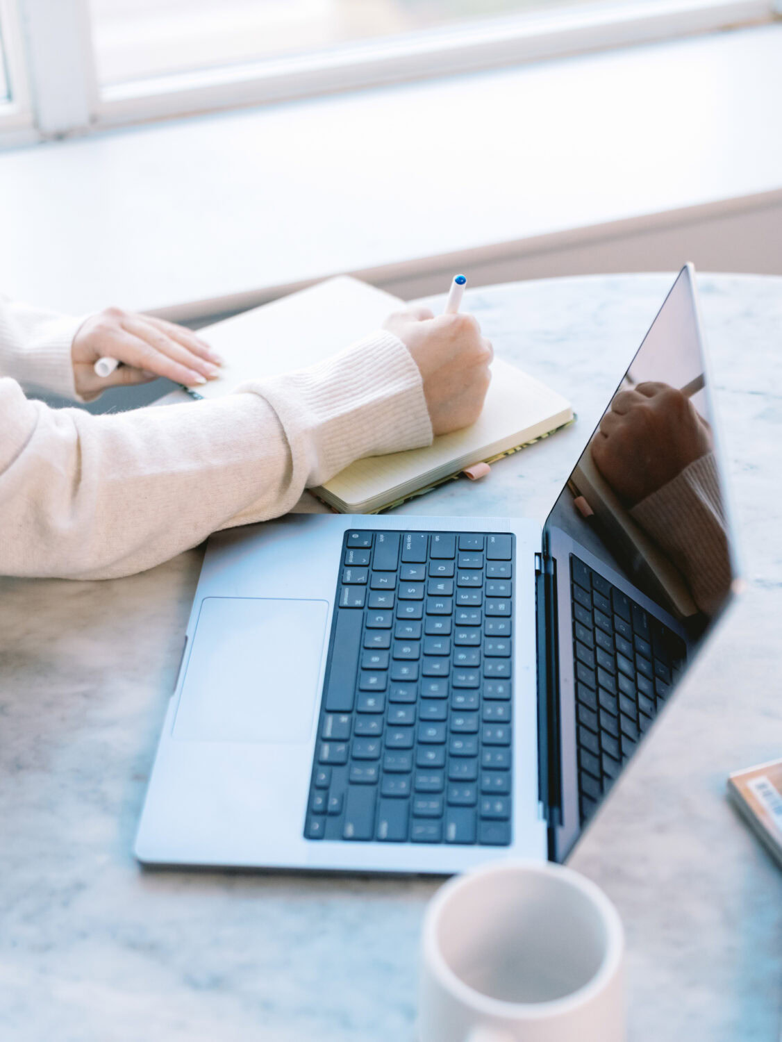 person in white sweater writing in a notebook next to a laptop on a gray marble table next to a bright window