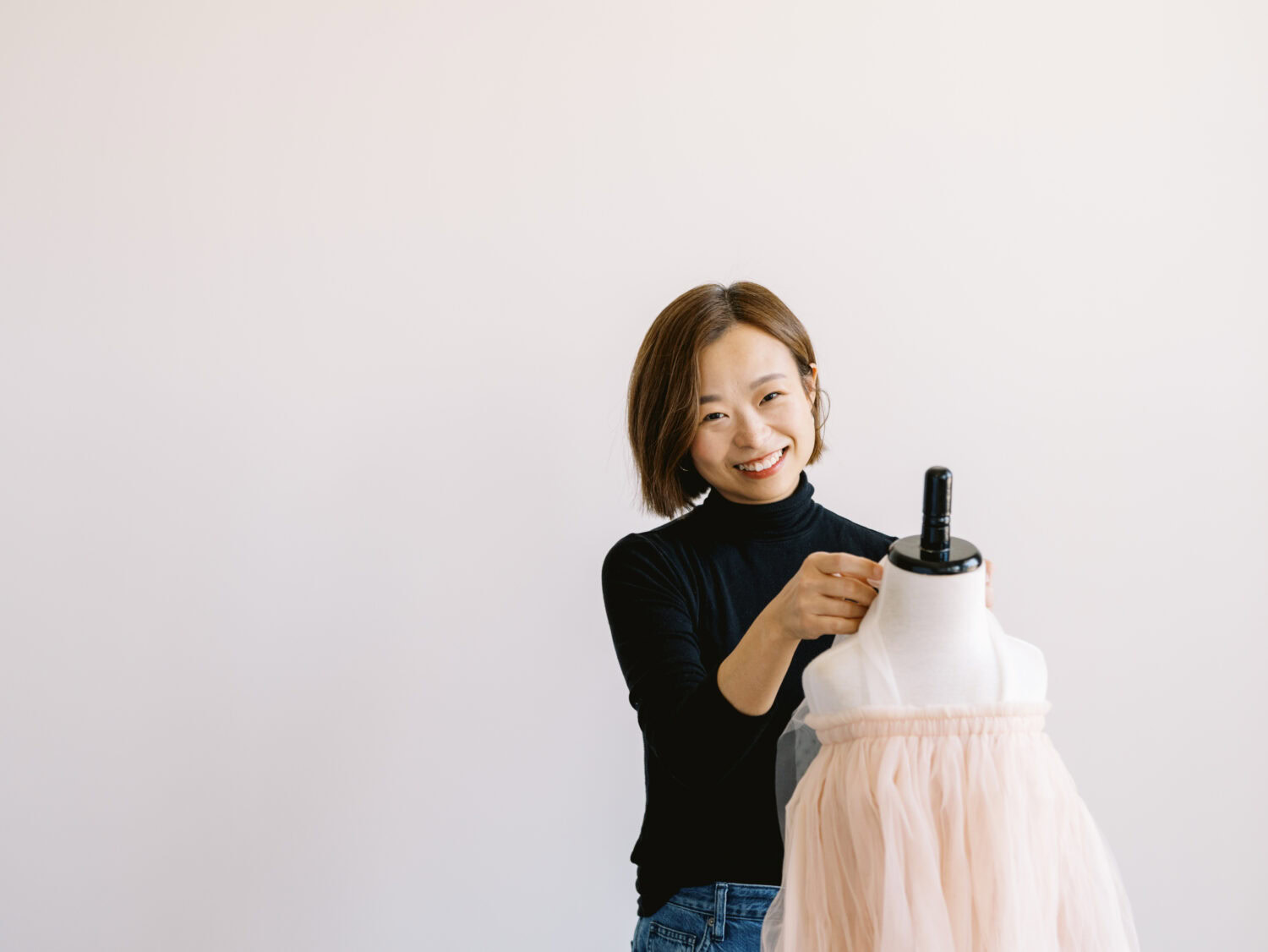 chinese designer in a black turtleneck fitting a pink tulle dress on a mannequin and smiling