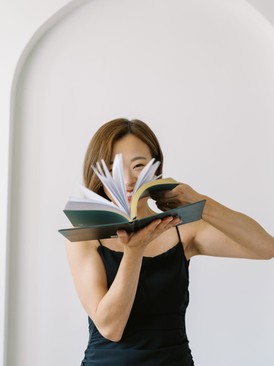woman in black dress smiling as she flips the pages of a book in front of her face