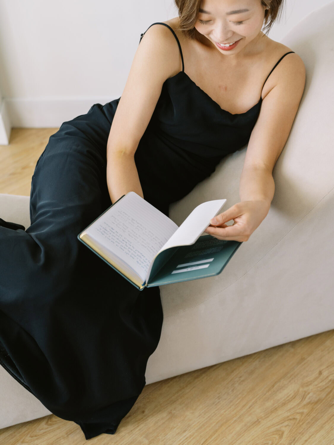 closeup of a woman in a black sleeveless dress sitting on a modern tan sofa reading a book